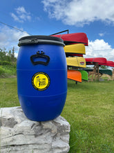 Load image into Gallery viewer, Blue food barrel with a yellow label on a stone ledge, with colorful canoes in the background.