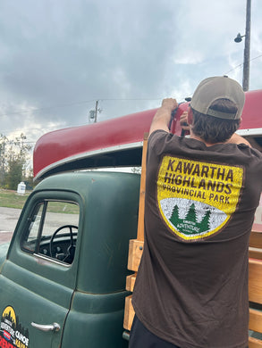 Person loading a canoe onto an old green truck with Kawartha Highlands Provincial Park t-shirt.