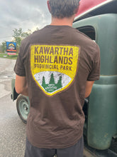 Load image into Gallery viewer, Person wearing a brown t-shirt with Kawartha Highlands Provincial Park logo, standing next to a green truck.
